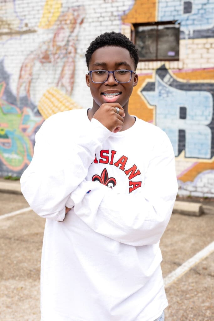 High school senior portrait of Black male student wearing glasses and white sweatshirt posing in front of mural in Baton Rouge